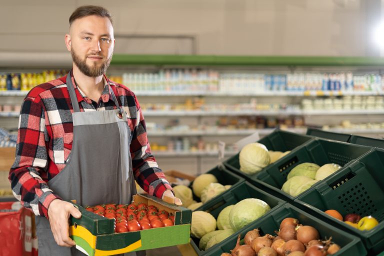 portrait of joyful male supermarket worker holding in hands box with fresh organic vegetables, looking at camera and smiling. Young man with organic